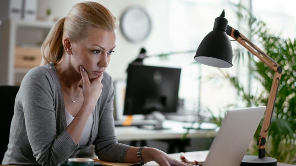 A focused blonde woman is sitting at an office desk, looking intently at her laptop screen with her hand on her chin.