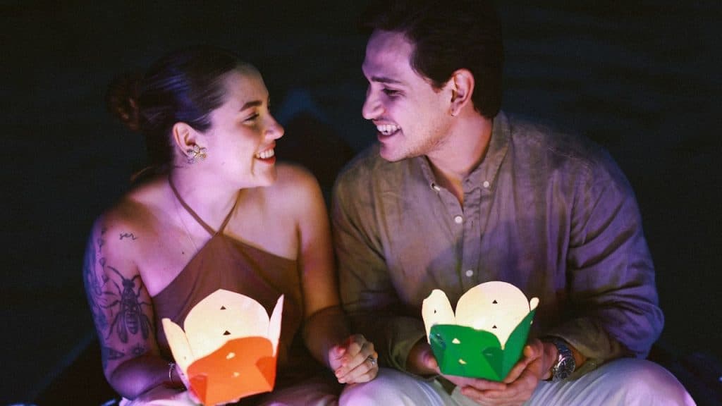 A couple is sitting in the dark, smiling and looking at each other while holding illuminated paper lanterns.