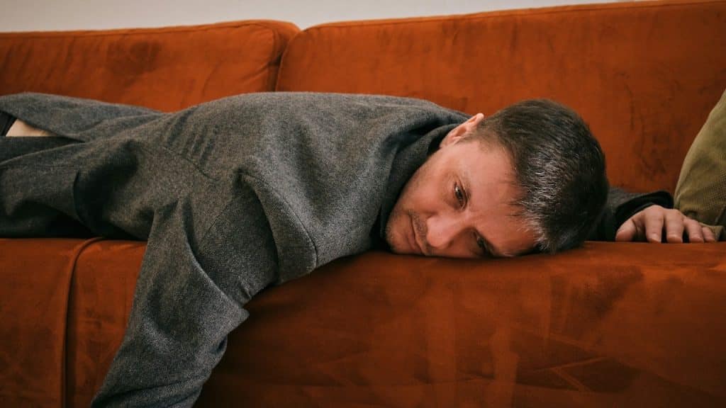 A man in a grey coat is lying face down on an orange couch, looking distressed.