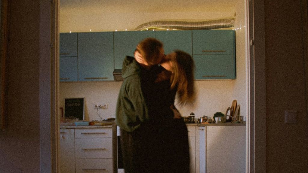 A couple is kissing and embracing in a dimly lit kitchen doorway.