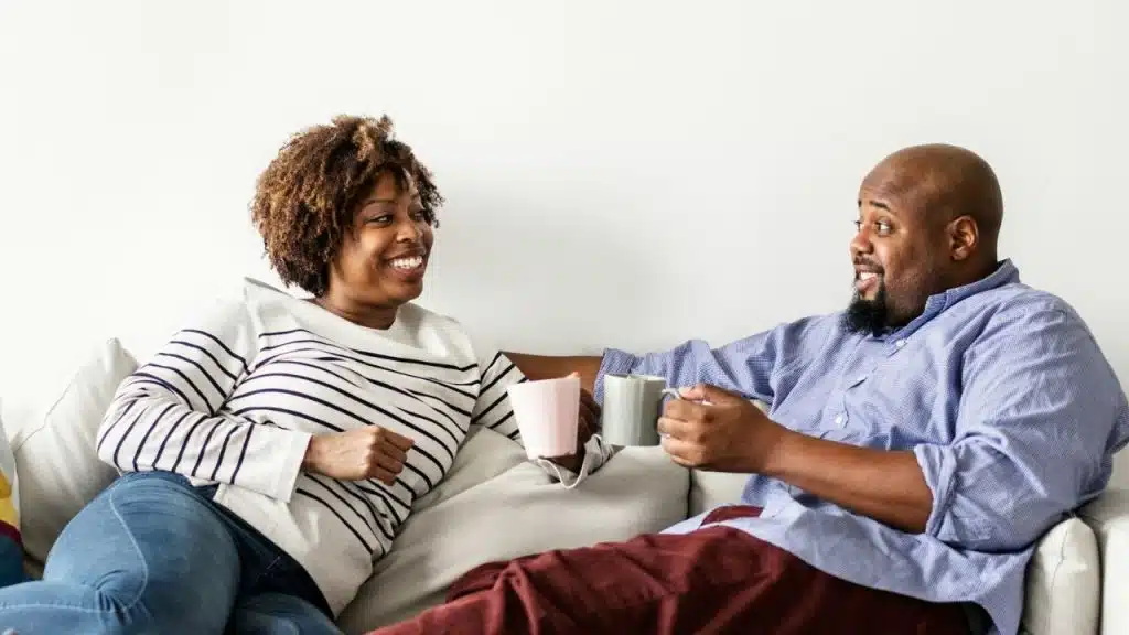 A man and woman sit on a couch, holding mugs and smiling at each other.
