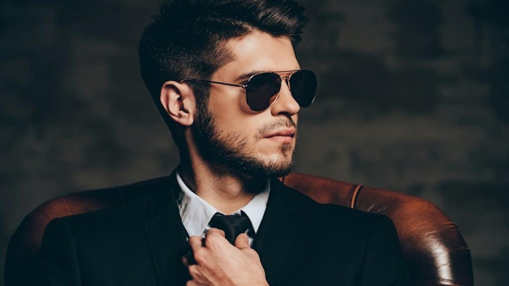 A man wearing sunglasses and a suit is adjusting his tie while looking to the side.