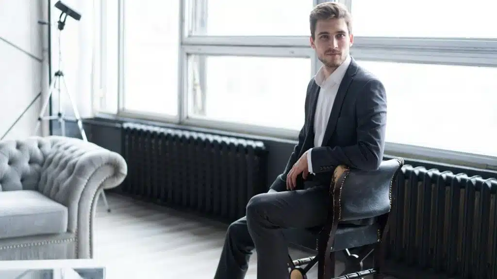 A confident, thoughtful young man in a full suit looking away while sitting on the stool.