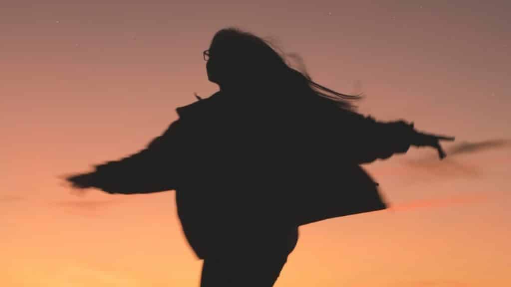 A silhouette of a person with long hair dancing or twirling against a vibrant sunset sky filled with stars and a crescent moon.
