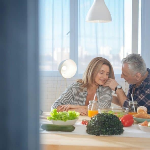 An older man affectionately touches a woman's face as they sit together at a dining table with fresh food in a sunlit room.