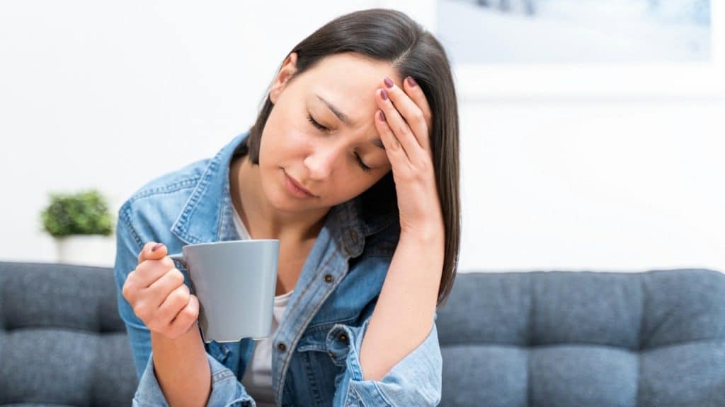 A woman with dark hair and a denim shirt sits on a blue couch, holding a gray mug while resting her head in her hand with a weary expression.