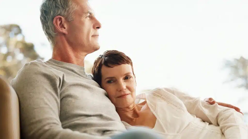 A content older couple sits together, with the woman leaning her head on the man's chest as they both look off-camera.