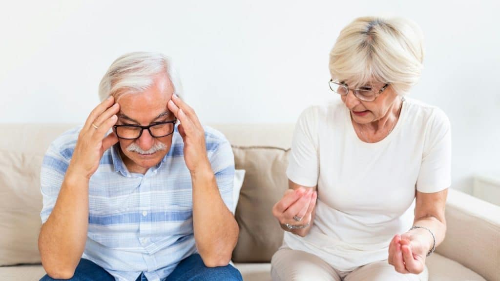 An older man in a blue-striped shirt sits on a couch with his head in his hands, while an older woman beside him looks on with an expressive, questioning gesture.