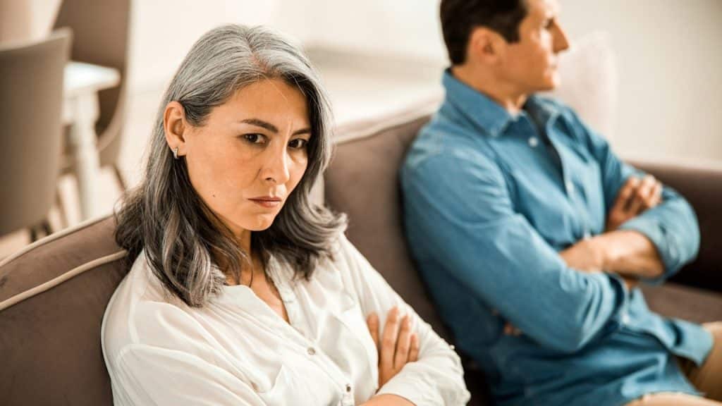 A woman with long gray hair with a stern expression and crossed arms, while a man sits turned away from her in the background on a couch.