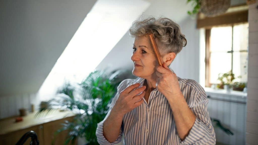 An older woman with short gray hair and a blue-and-white striped shirt smiles while combing her hair in a bright room filled with plants.