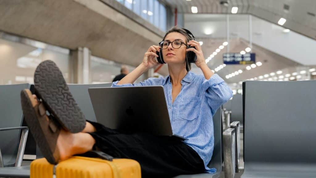A woman in a blue-striped shirt and glasses sits in an airport terminal, wearing large headphones and looking ahead thoughtfully while resting her feet on a yellow suitcase.