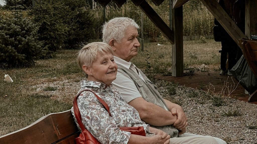An older couple sits side-by-side on a wooden bench outdoors; the woman smiles warmly while the man maintains a thoughtful, neutral expression.