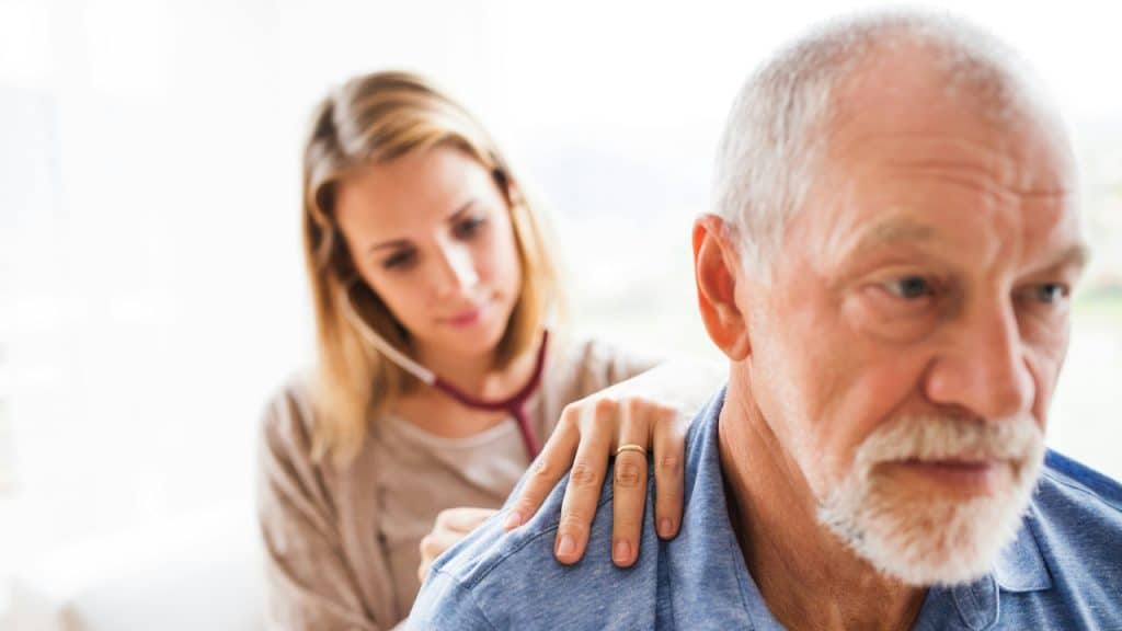 A woman using a stethoscope to check an old man’s heartbeat while placing it on his back.