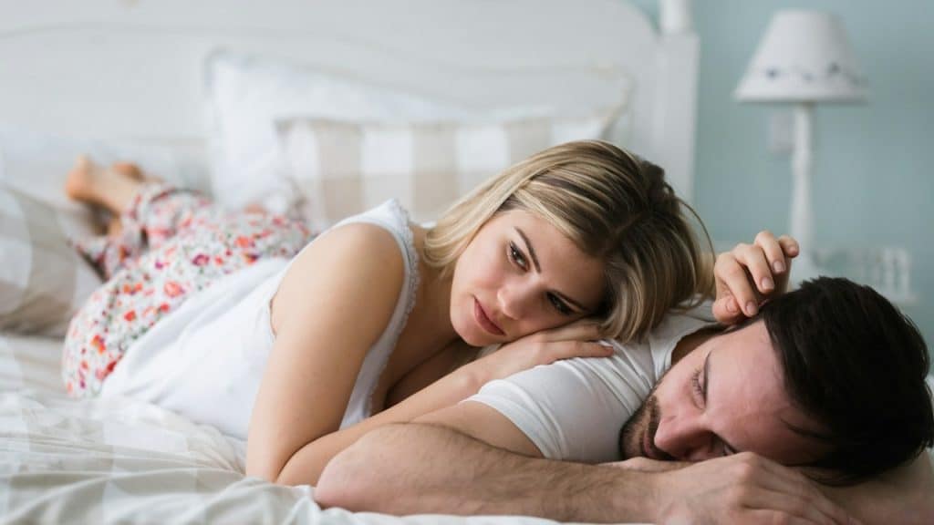 A young woman lying on a bed with her husband, who is sleeping.