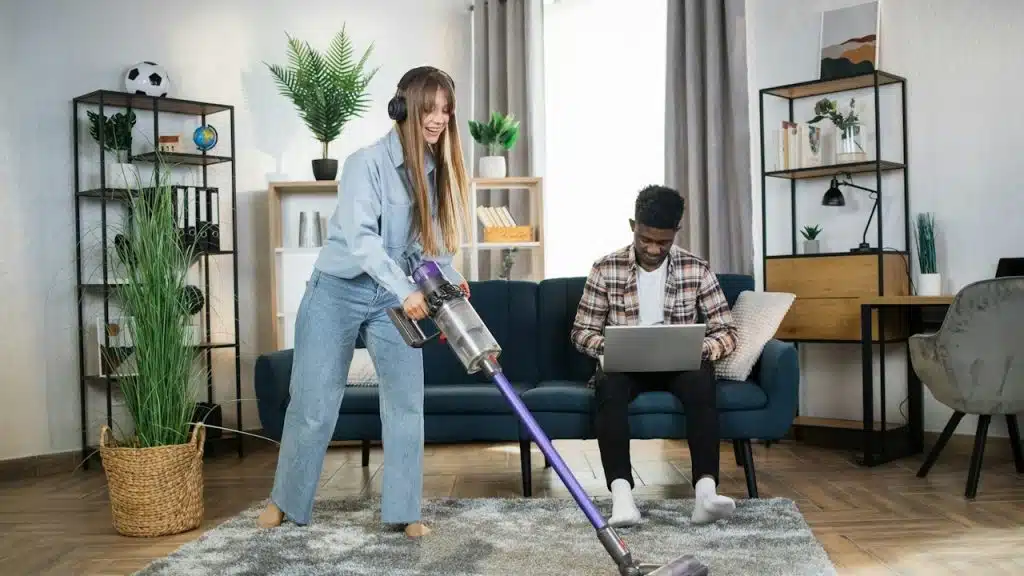 A woman wearing headphones cleaning the carpet while her husband is working on a laptop.
