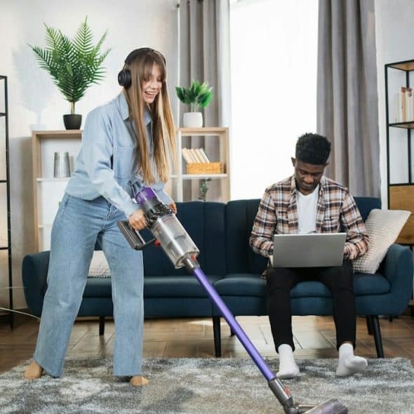 A woman wearing headphones cleaning the carpet while her husband is working on a laptop.