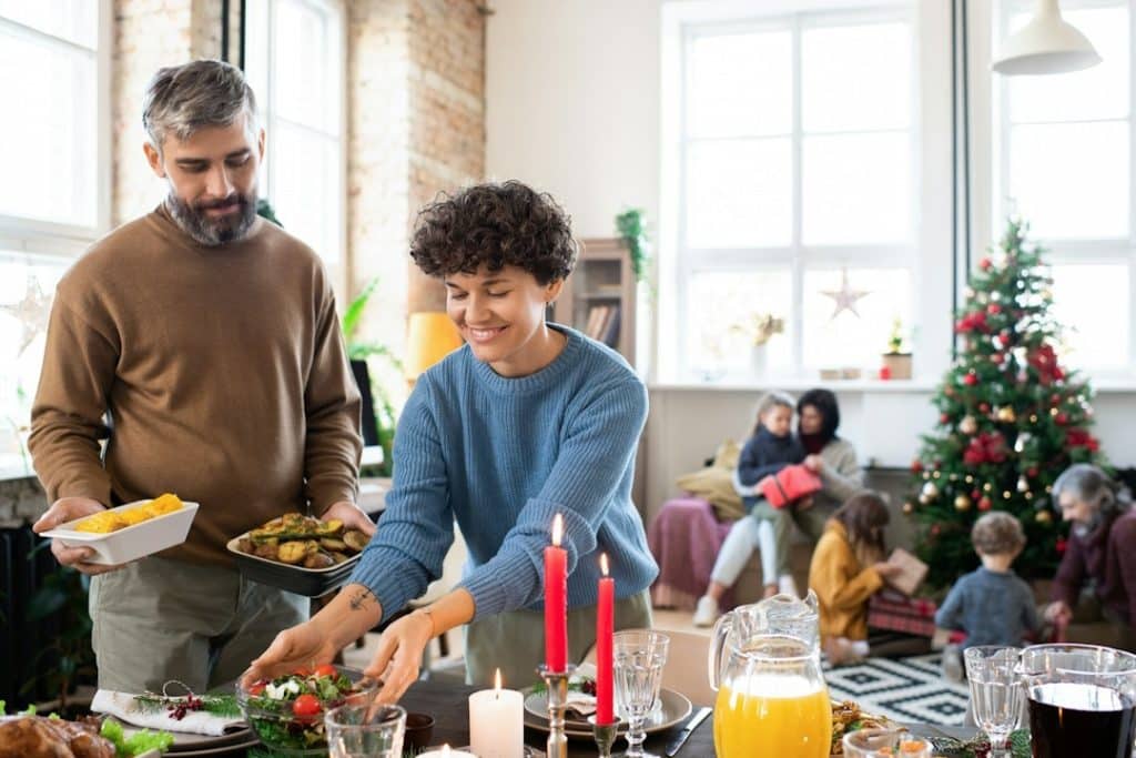 A happy husband and wife putting homemade salad and baked potatoes, drinks, and other food on a served festive table before a family Christmas dinner.