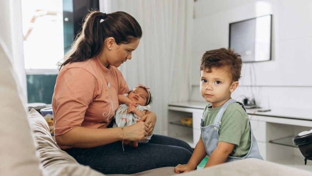 A woman is sitting on a couch holding a baby while her toddler is standing next to her.