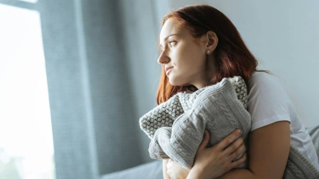 A woman with red hair looks out a window while hugging a thick grey sweater.