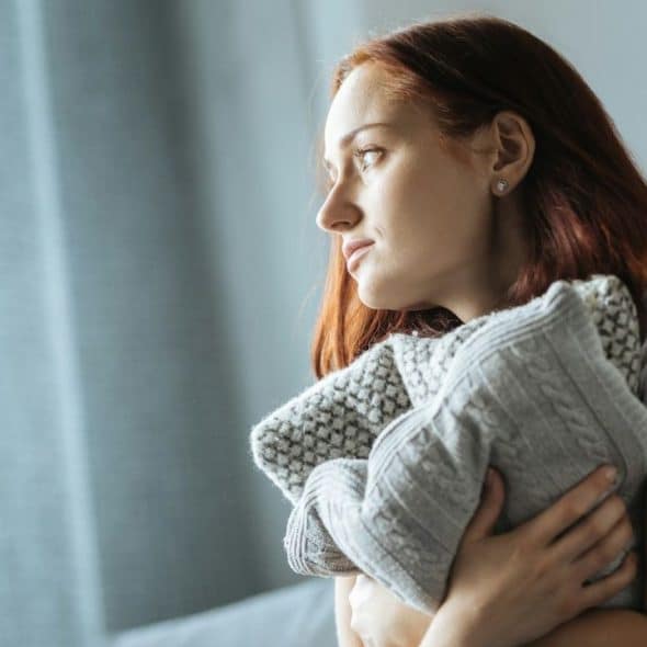 A woman with red hair looks out a window while hugging a thick grey sweater.