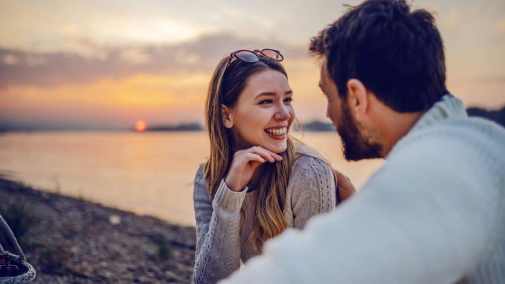 A smiling woman looks at a man's face during sunset by a body of water.