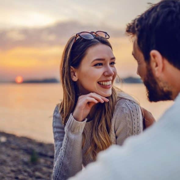 A smiling woman looks at a man's face during sunset by a body of water.