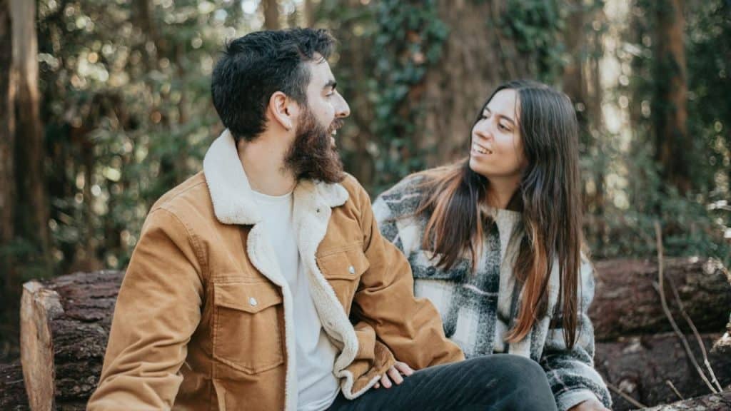 A man and woman sit on a log in a forest, looking at each other.