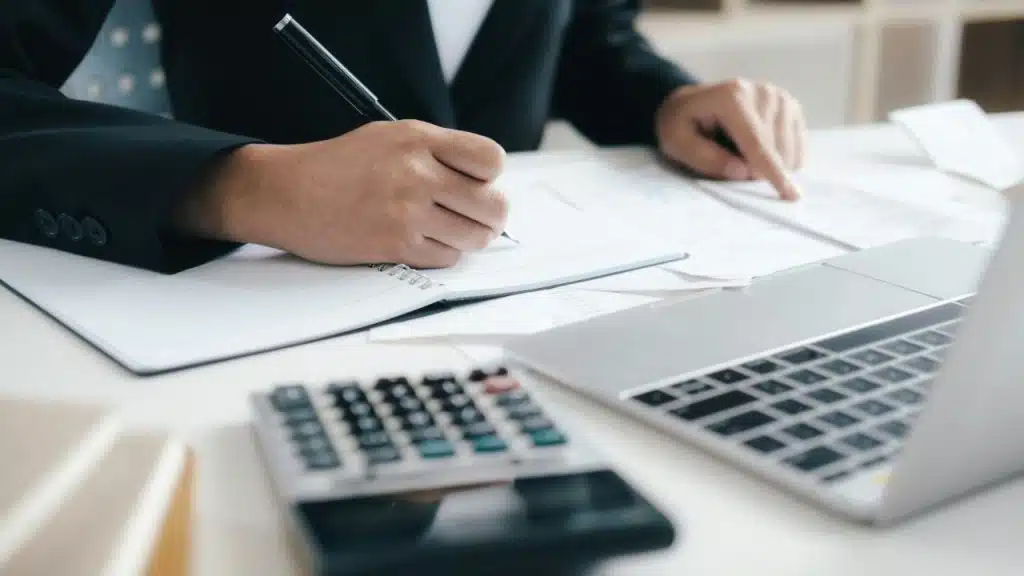 Hands in a suit write on paper with a calculator and laptop on a desk.