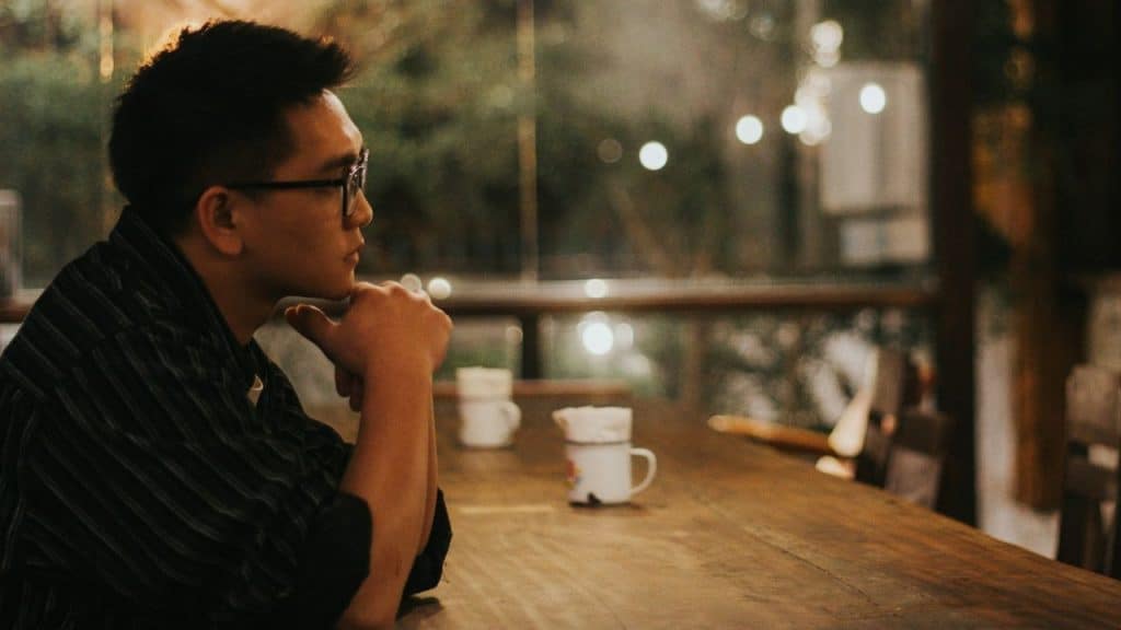 A man with glasses rests his chin on his hand at a long wooden table.