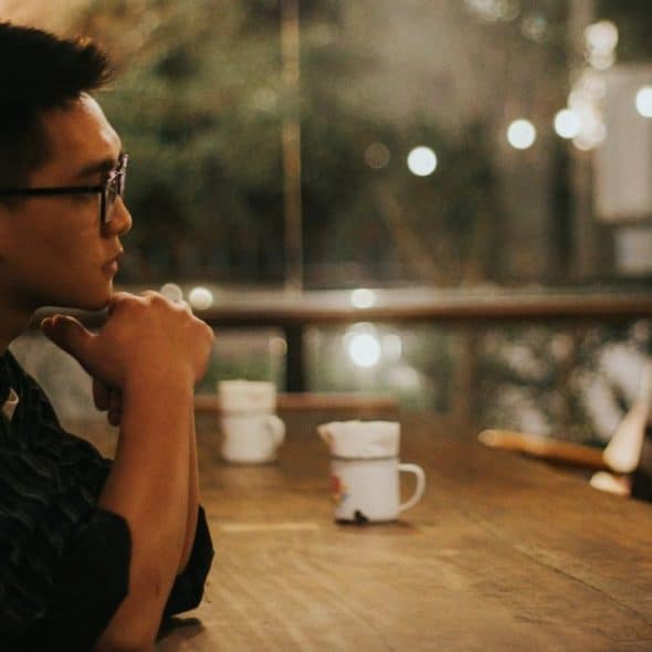 A man with glasses rests his chin on his hand at a long wooden table.