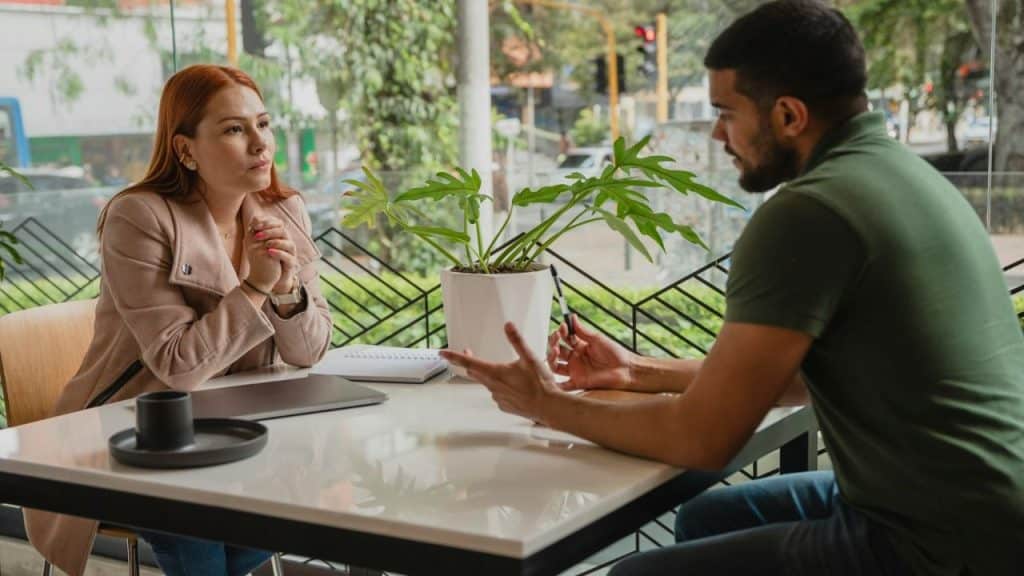 A man and woman sit at a white table talking near a large window.