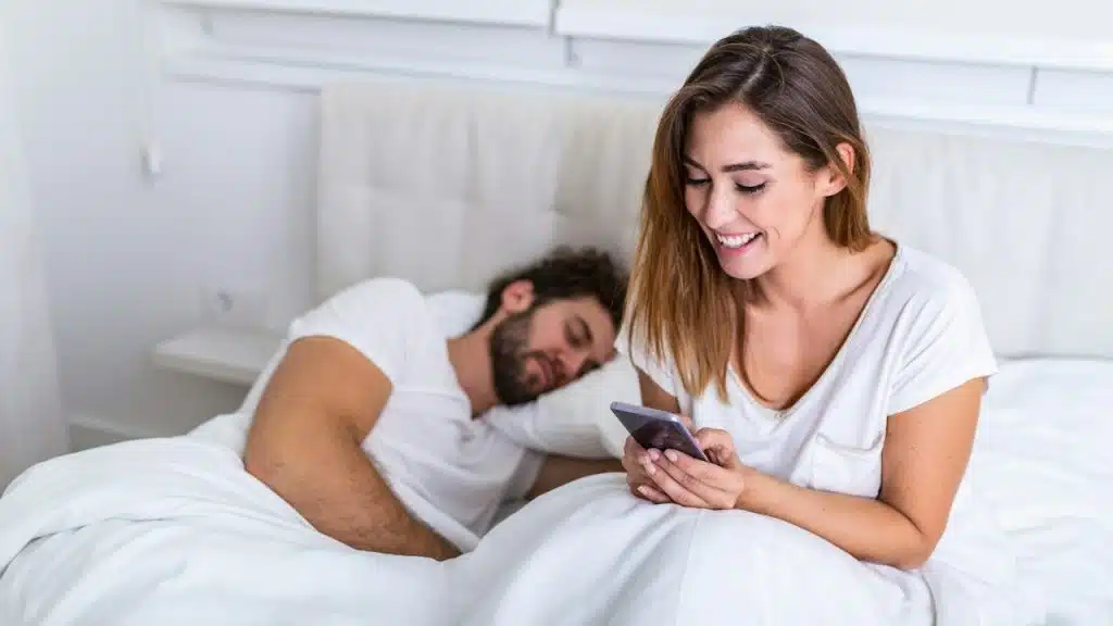 A beautiful woman talking privately on the phone while her husband is sleeping in the bed.