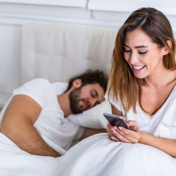 A beautiful woman talking privately on the phone while her husband is sleeping in the bed.