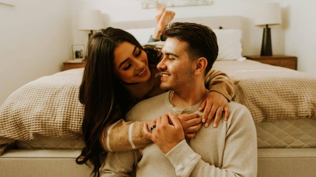 A woman lying on the bed is hugging her husband from behind, who is sitting on the floor.
