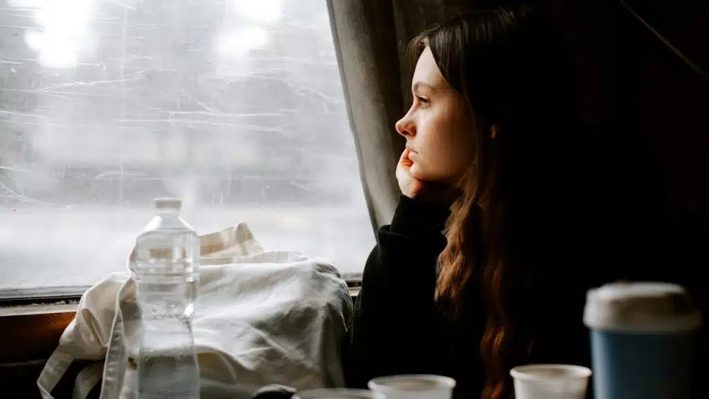 A woman sitting by a window and staring out of it while she sits at a table on which paper cups and a water bottle are placed next to her bag.