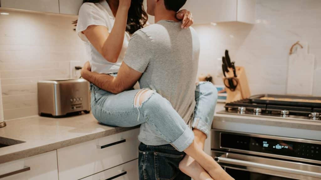 A couple embraces in a modern kitchen while the woman sits on the counter.