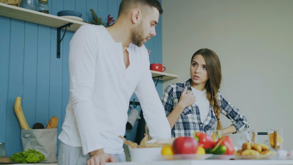 A man and woman engage in a serious conversation in a kitchen, surrounded by fresh food.