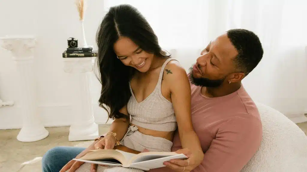 A happy couple sits together reading a book in a bright room.