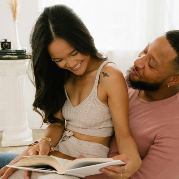 A happy couple sits together reading a book in a bright room.