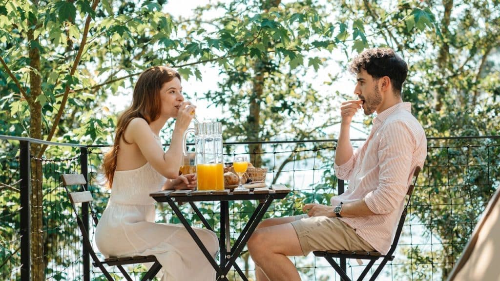 A man and woman share a meal at a small table on a sunlit wooden deck, surrounded by lush trees.