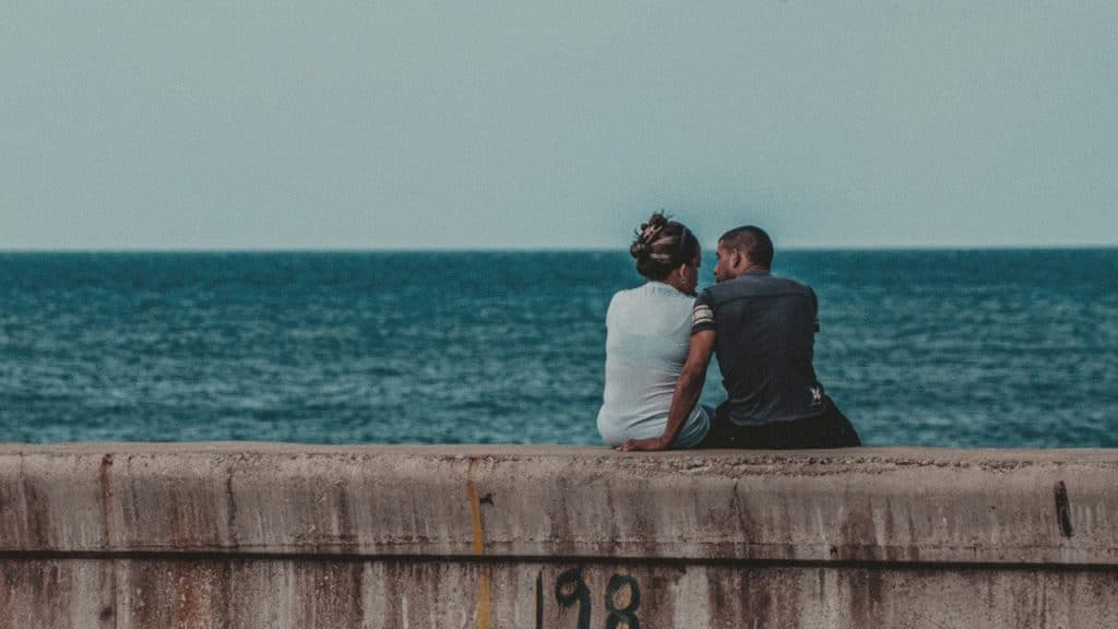 A man and woman sit together on a concrete wall, facing each other with the expansive ocean in the background.