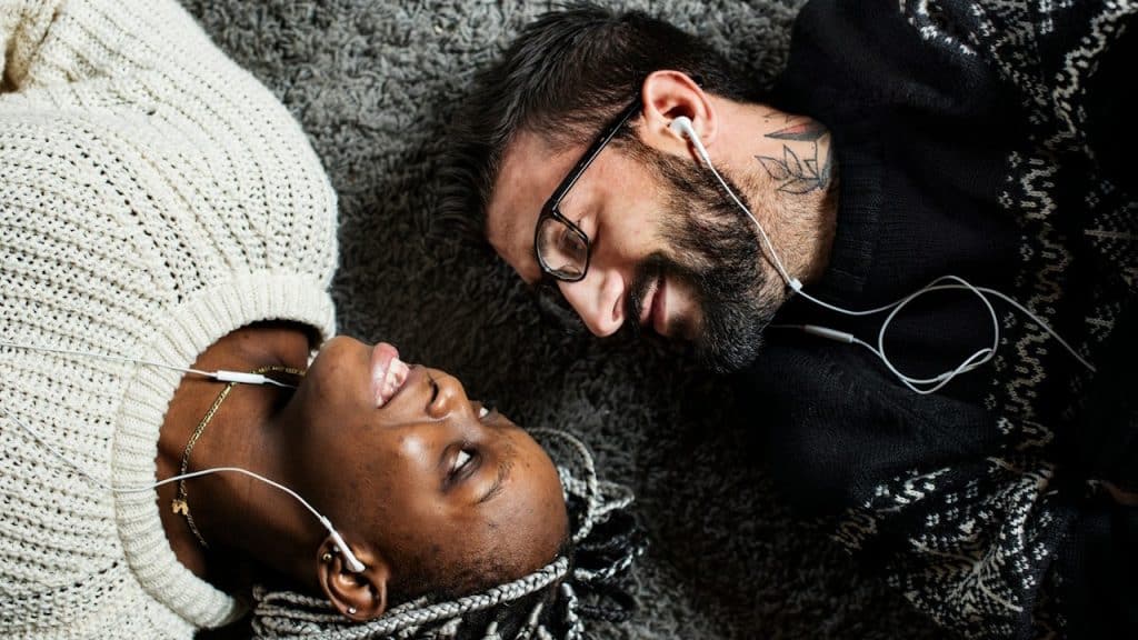 A smiling man and woman lie on a rug, facing each other while sharing wired earphones.