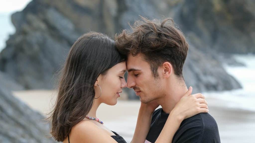 A young couple embraces on a beach with their eyes closed and foreheads touching, set against a backdrop of large coastal rock formations.
