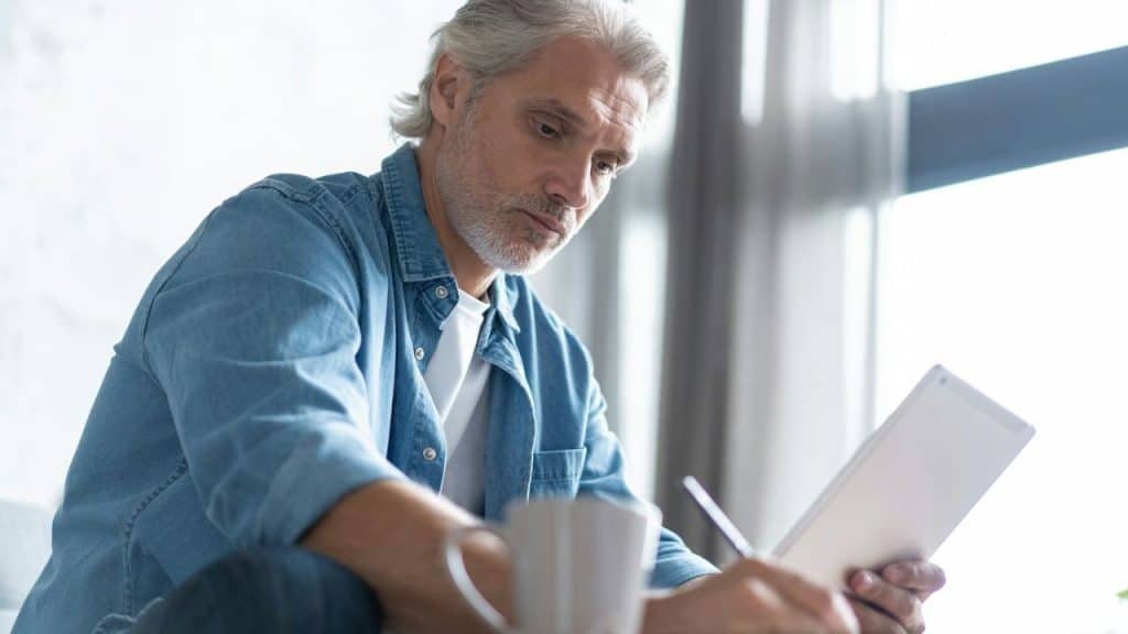 A middle-aged man in a denim shirt writes on a tablet next to a white mug.