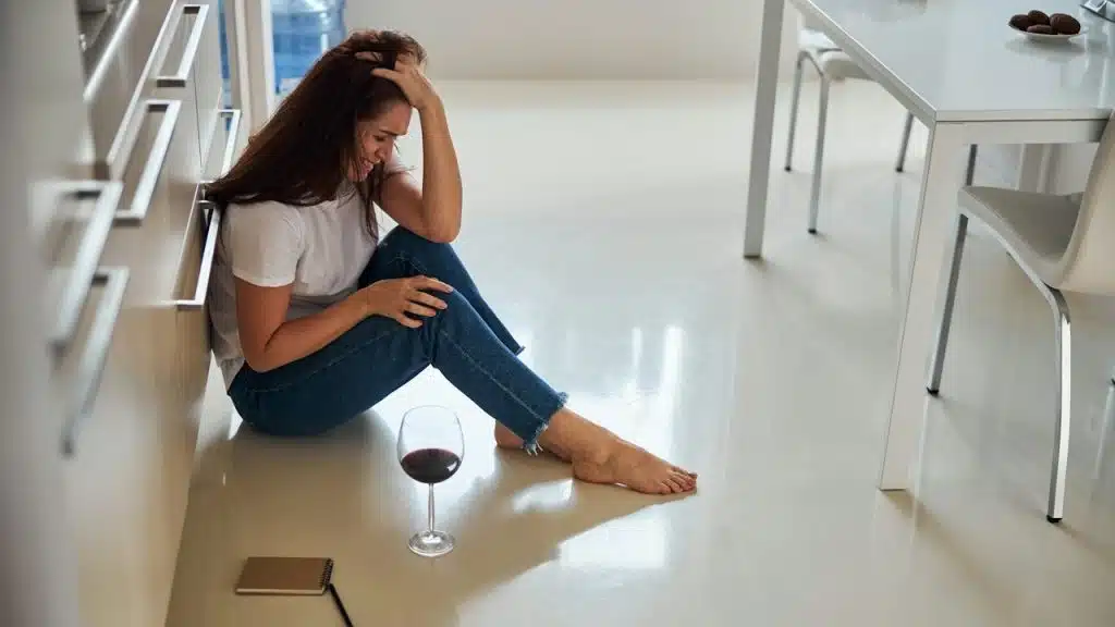 A crying woman is sitting on a floor with glass of wine.