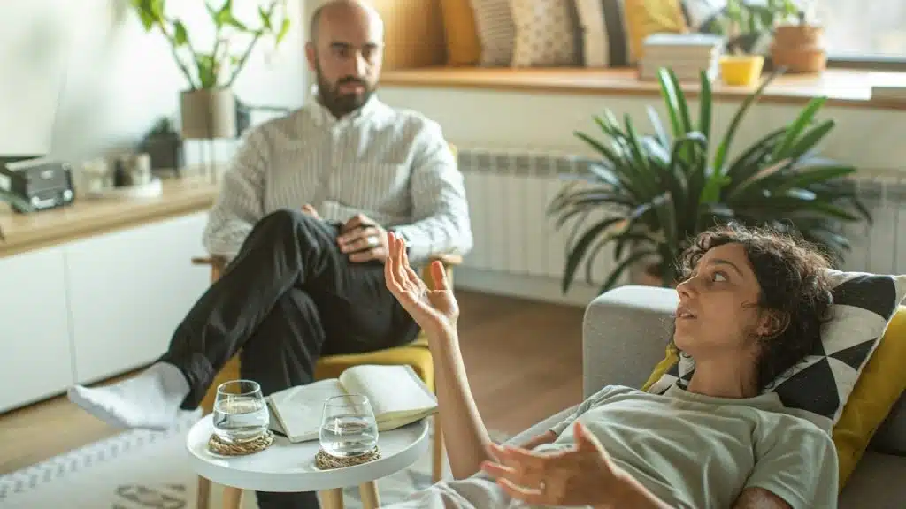 A woman laying on a couch is talking to a man sitting in a chair near her.