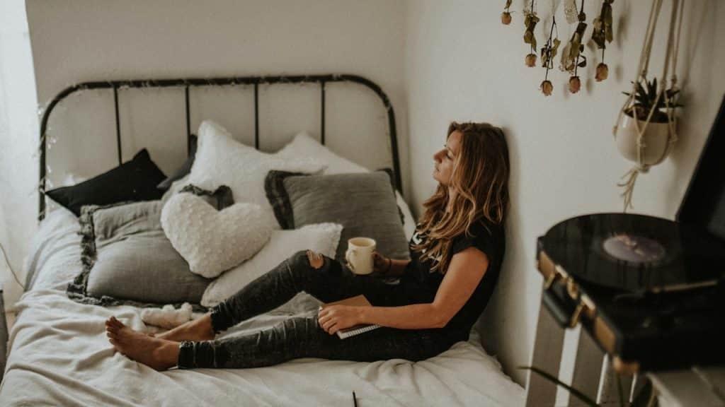 A woman sitting on a bed, leaning against the wall, holding a mug and a notebook.