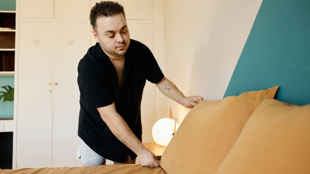 A man in a black shirt makes a bed with brown linens in a bedroom.
