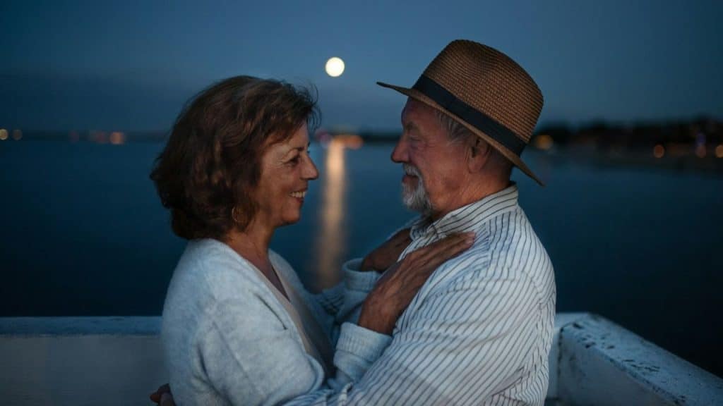 Older couple embracing outdoors at night with a full moon reflecting on the water.