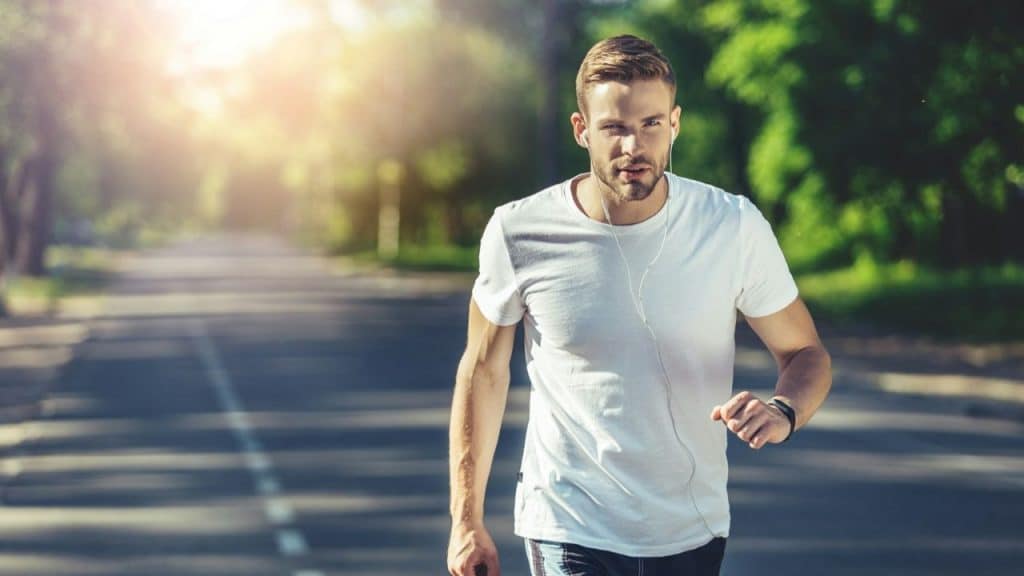 Man in a white T-shirt walking or running on a sunny, tree-lined road.